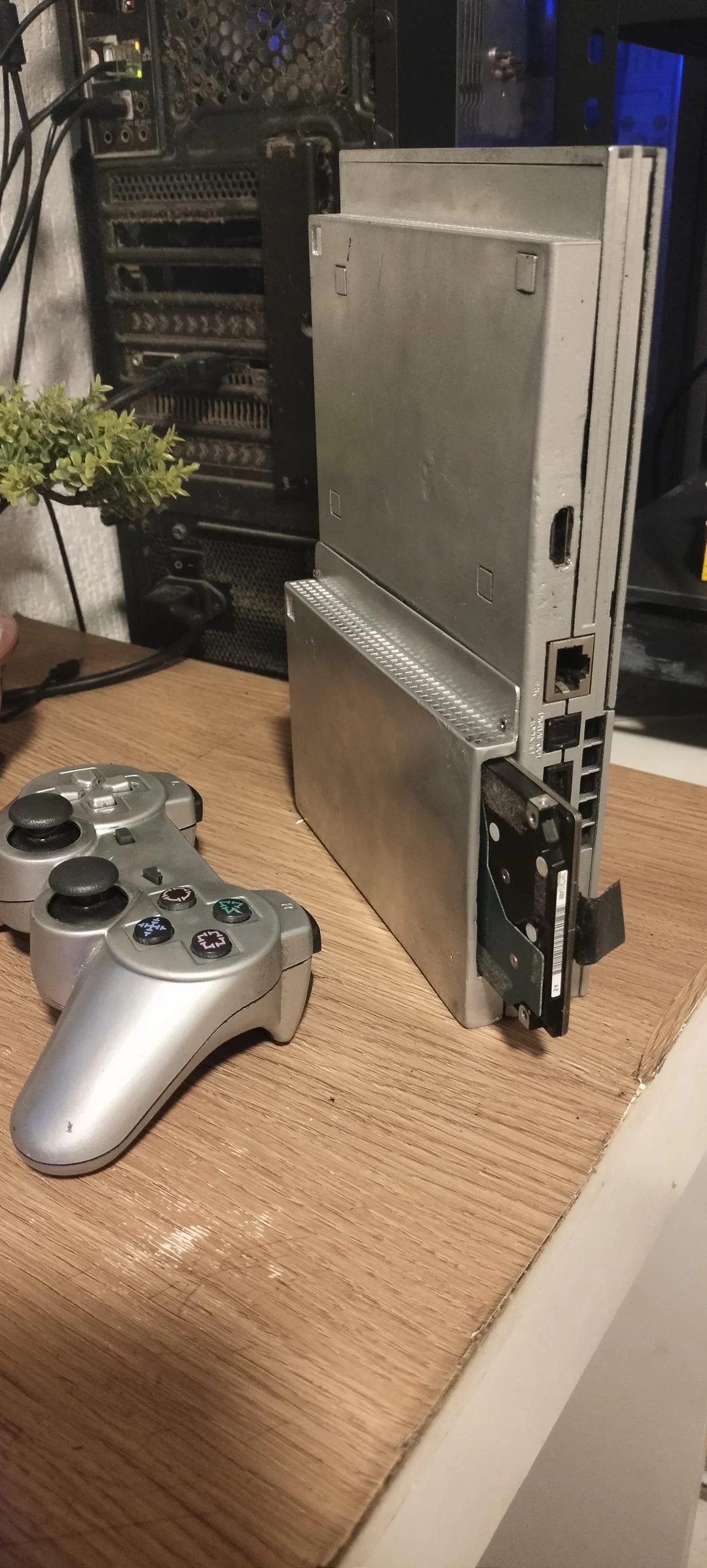 PlayStation 2 slim modificado com HD interno exposto, porta HDMI e controle DualShock prateado sobre uma mesa de madeira