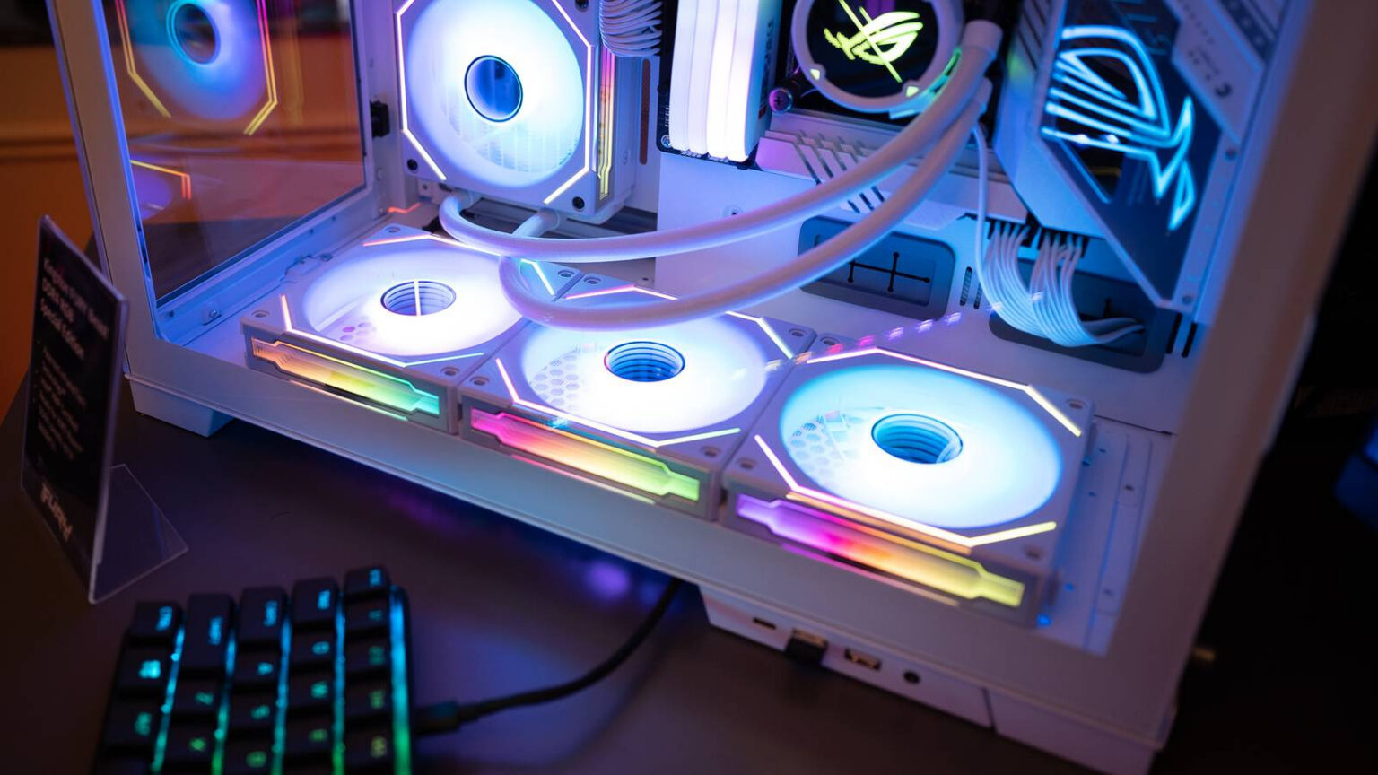 Gabinete de PC branco com ventoinhas de iluminação RGB, tubos de water cooler e teclado gamer iluminado em primeiro plano