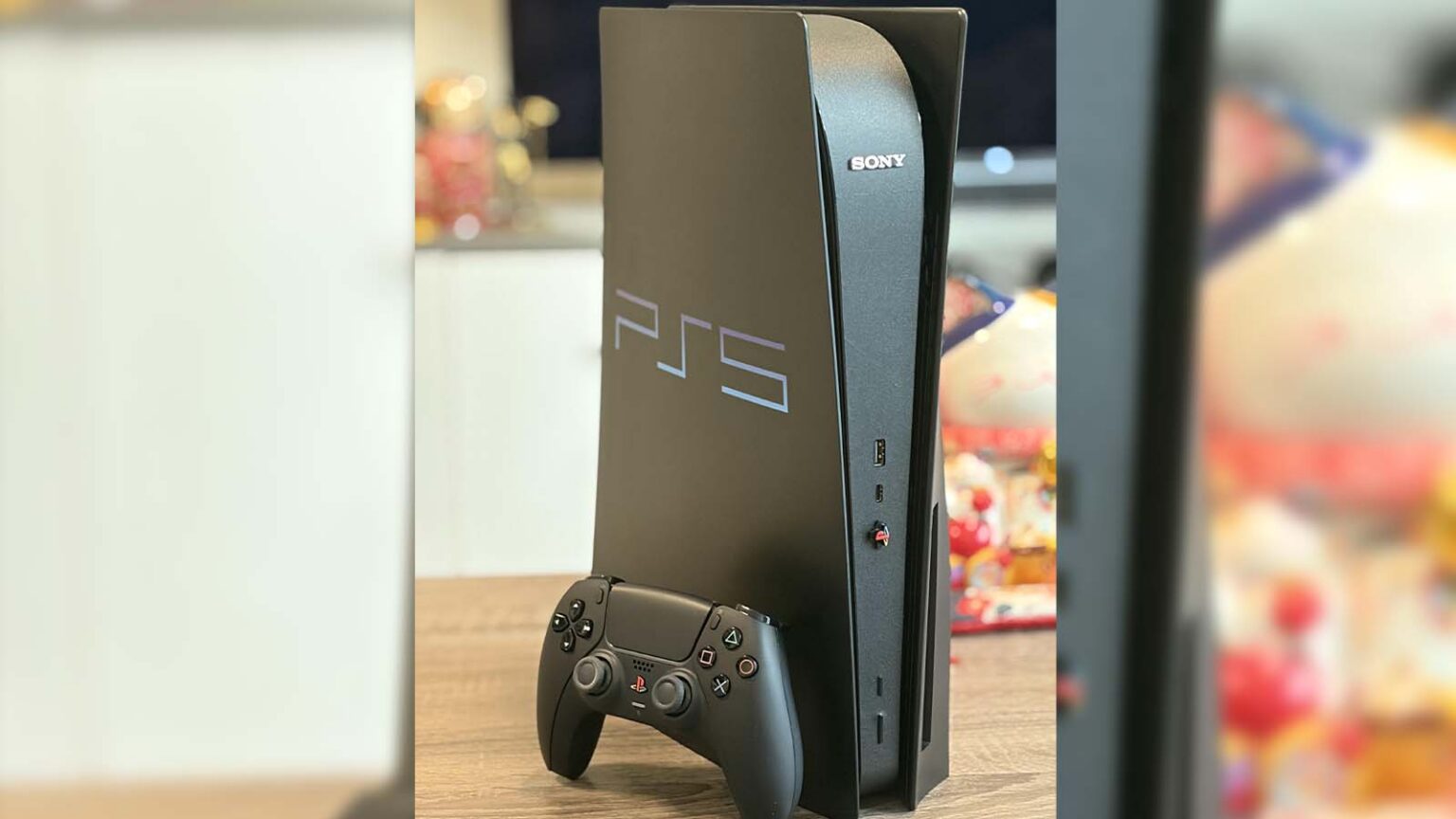 Console PlayStation 5 preto na vertical com logo PS5 e marca Sony, ao lado de um controle DualSense sobre uma mesa de madeira