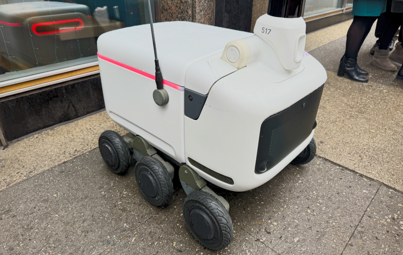 Robô de entrega branco com seis rodas, antena e número 517 no topo, estacionado em uma calçada ao lado de uma vitrine