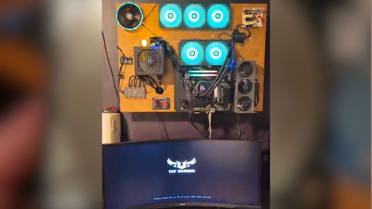 PC gamer montado em painel de madeira na parede, com ventoinhas RGB azuis, placa-mãe, fonte e placa de vídeo expostas, e monitor curvo com logo TUF Gaming