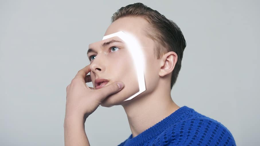 Homem jovem de suéter azul com a mão no queixo, olhando para cima, com uma moldura luminosa sobreposta no rosto simulando reconhecimento facial de IA