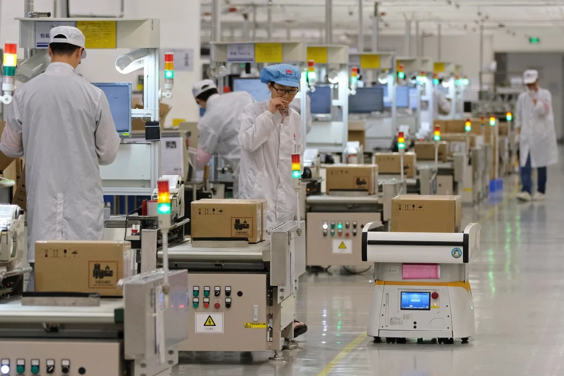 Linha de montagem de fábrica com trabalhadores de jaleco e robô autônomo transportando caixas de papelão entre bancadas de produção