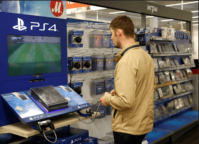 Homem jogando futebol em um PlayStation 4 exposto em loja de eletrônicos, com controle conectado ao console e prateleiras de jogos ao fundo