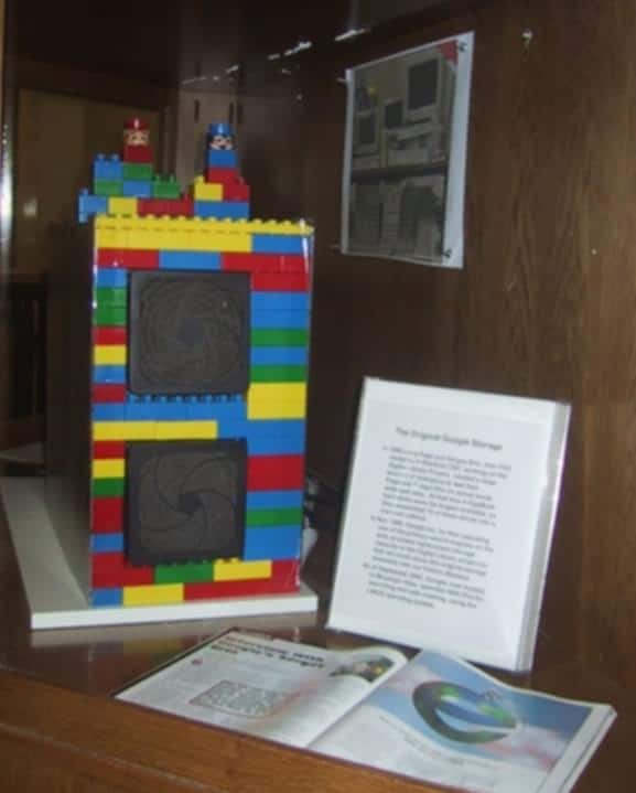 Maquete em LEGO de um dos primeiros servidores do Google, exposta em vitrine com placa explicativa e revista aberta ao lado