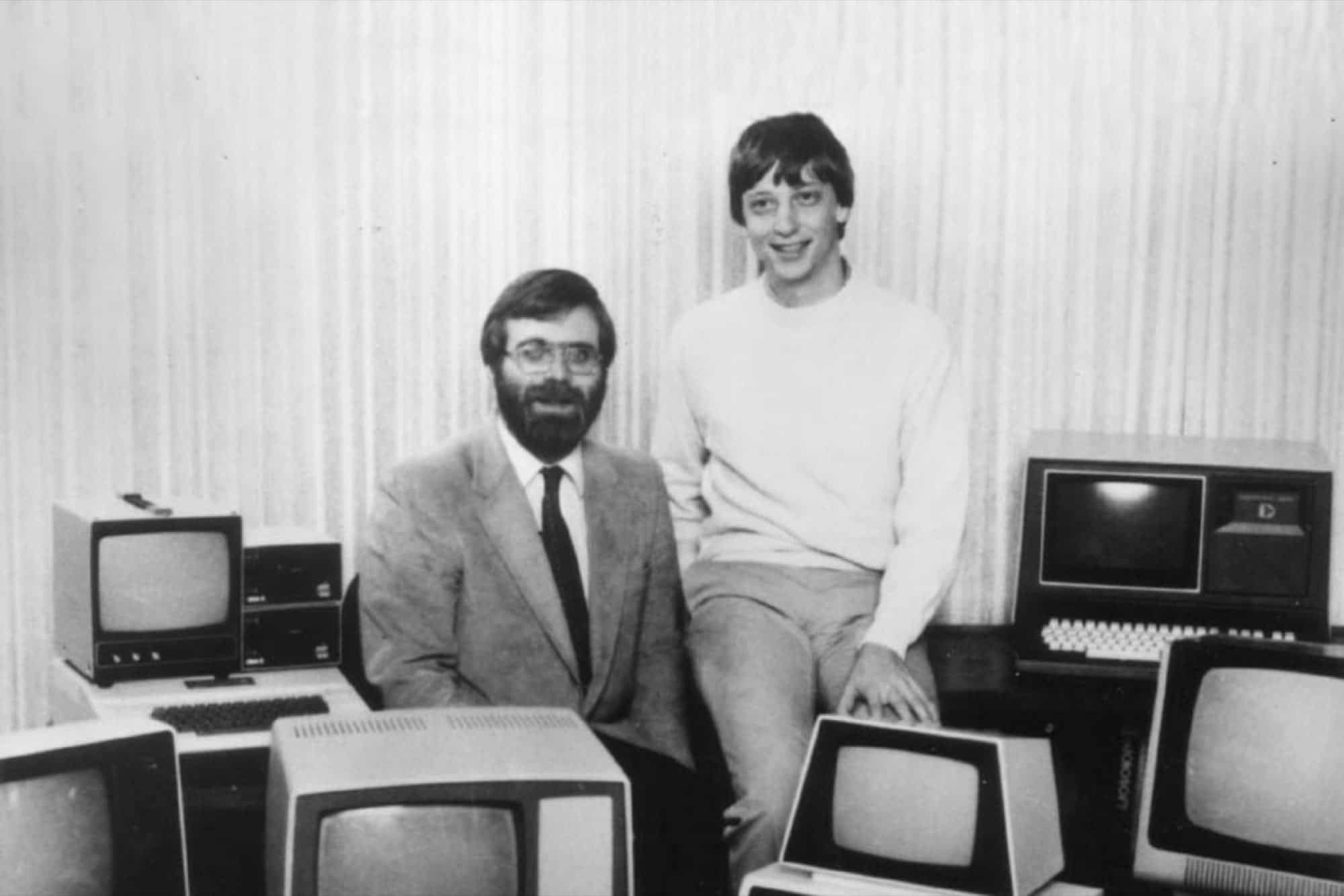 Foto em preto e branco de dois homens sentados entre antigos computadores de mesa com monitores de tubo, um de barba e óculos de terno, outro sorrindo de suéte…