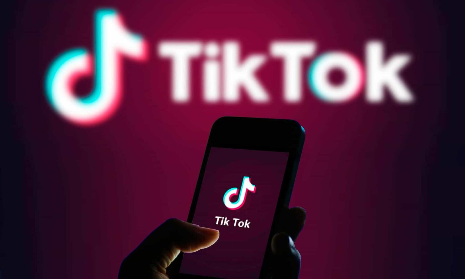 Smartphone com o logo do TikTok na tela, segurado por uma mão em silhueta, com o logo do TikTok desfocado ao fundo sobre fundo vinho