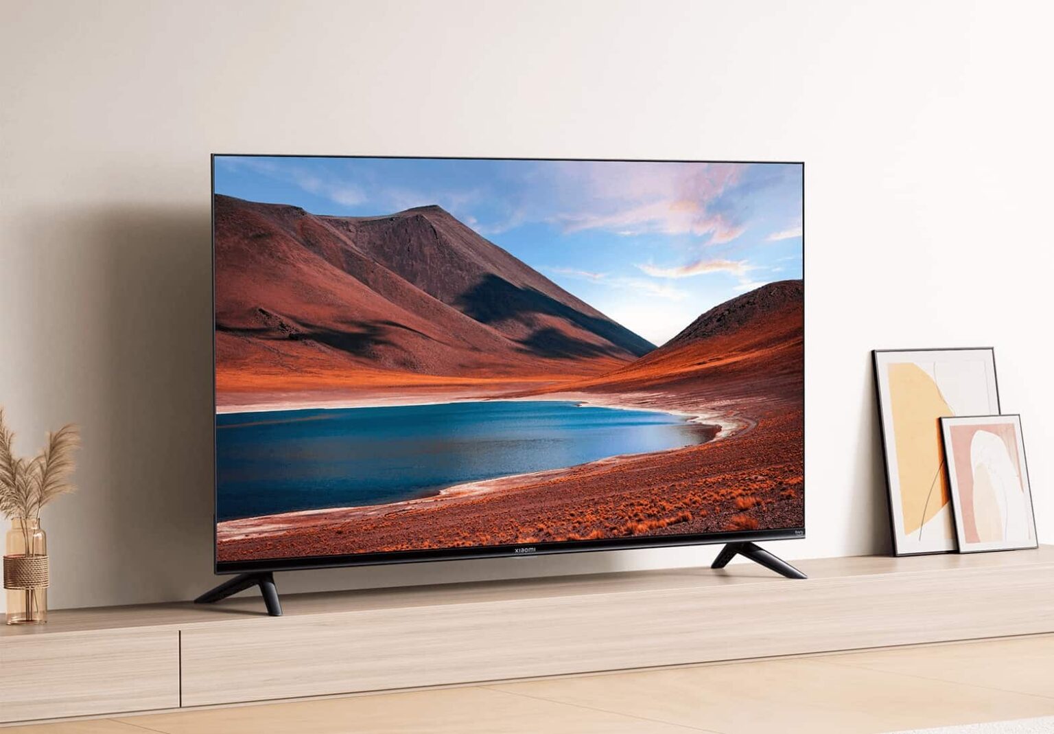 Smart TV Xiaomi de tela plana sobre móvel de madeira, exibindo paisagem desértica com lago azul e montanhas avermelhadas ao entardecer