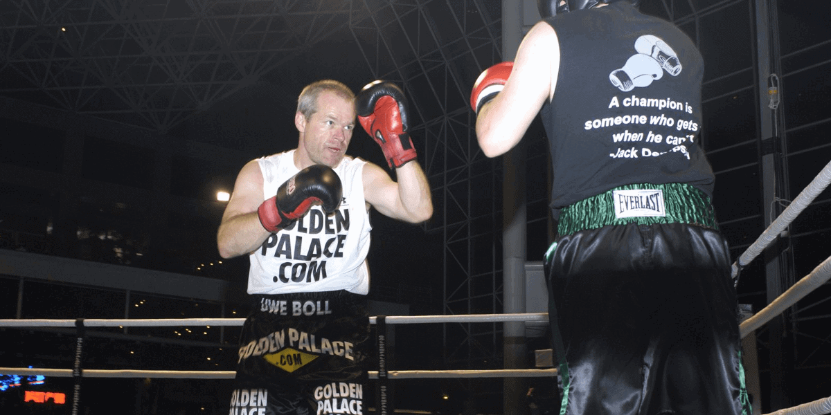 Dois homens treinando boxe no ringue, um deles de regata com a inscrição GOLDEN PALACE.COM e luvas vermelhas, enquanto o outro veste camiseta com a frase "A ch…