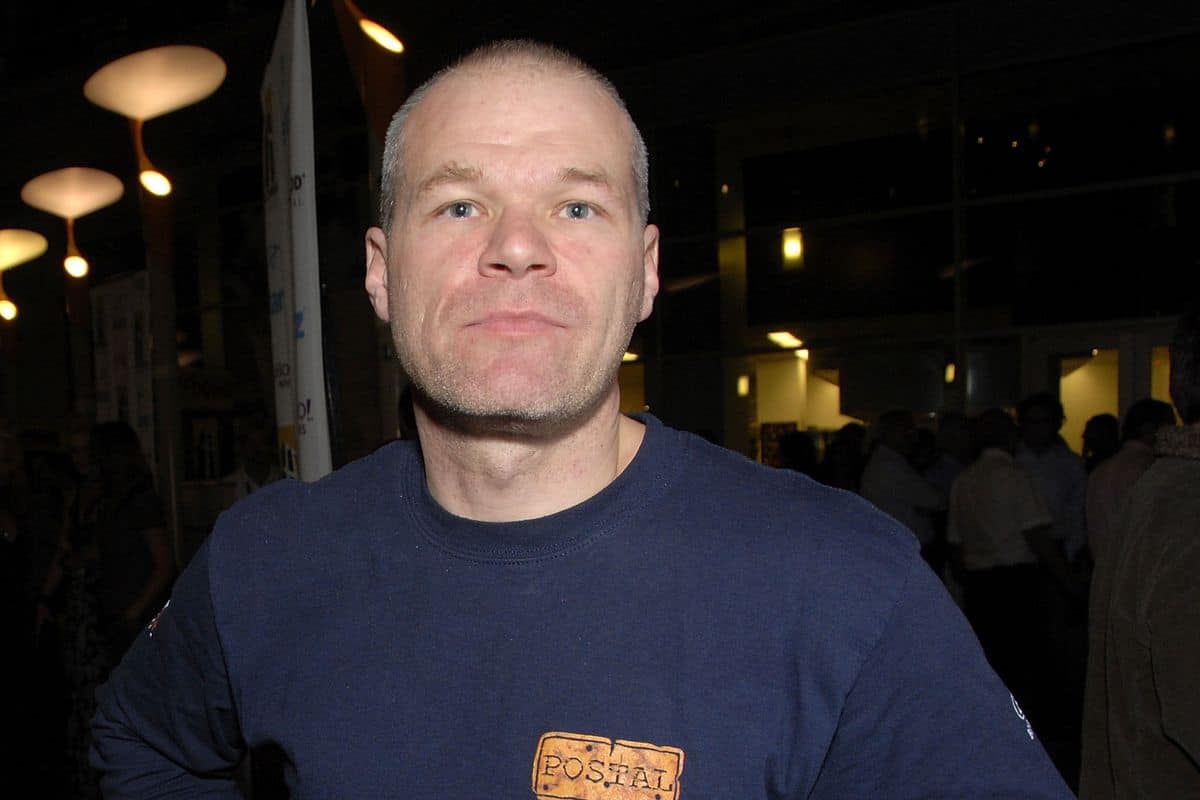 Homem careca de camiseta azul-marinho com estampa "POSTAL" em um evento com pessoas ao fundo