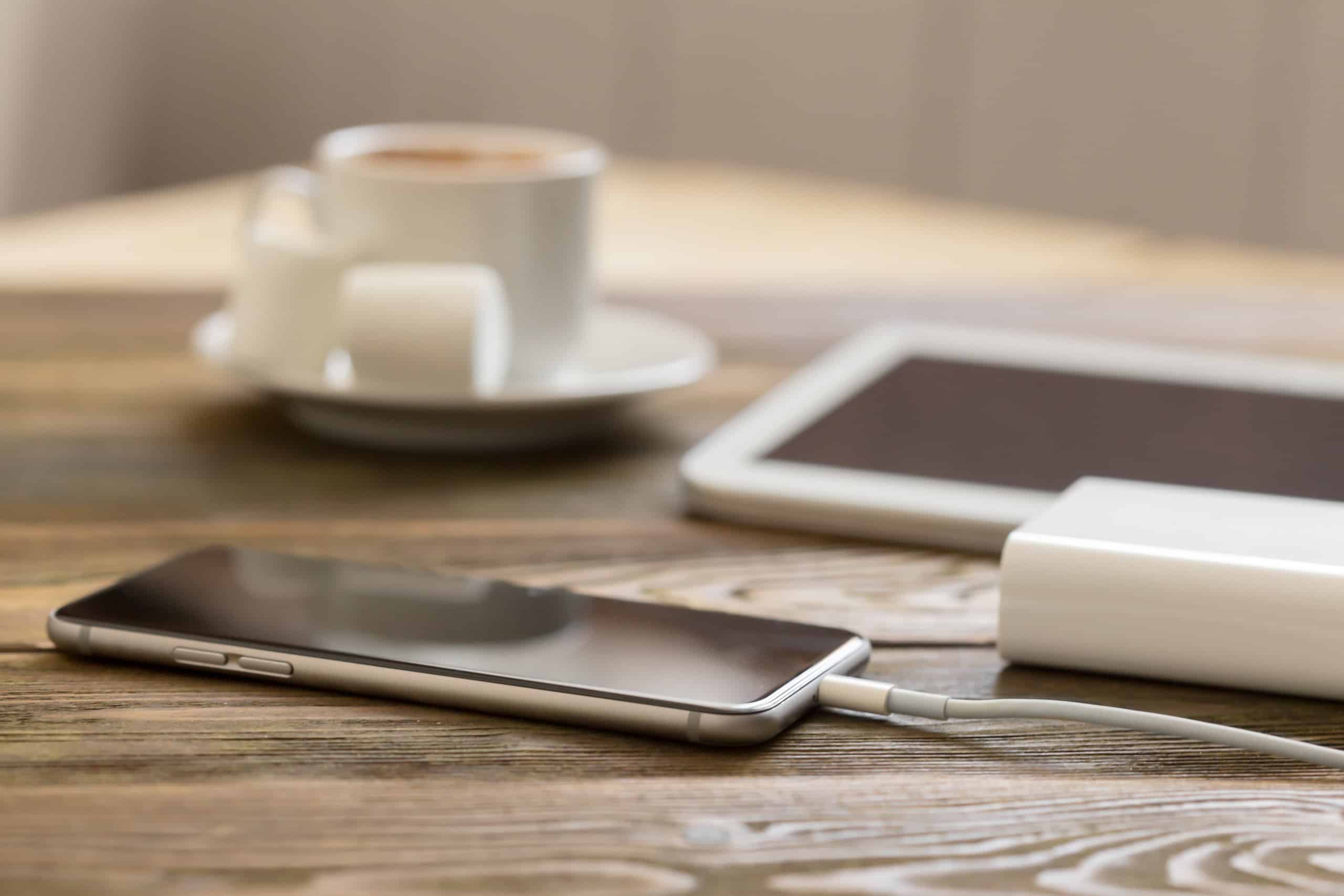 Smartphone branco carregando por cabo conectado a um power bank, ao lado de um tablet e uma xícara de café sobre mesa de madeira