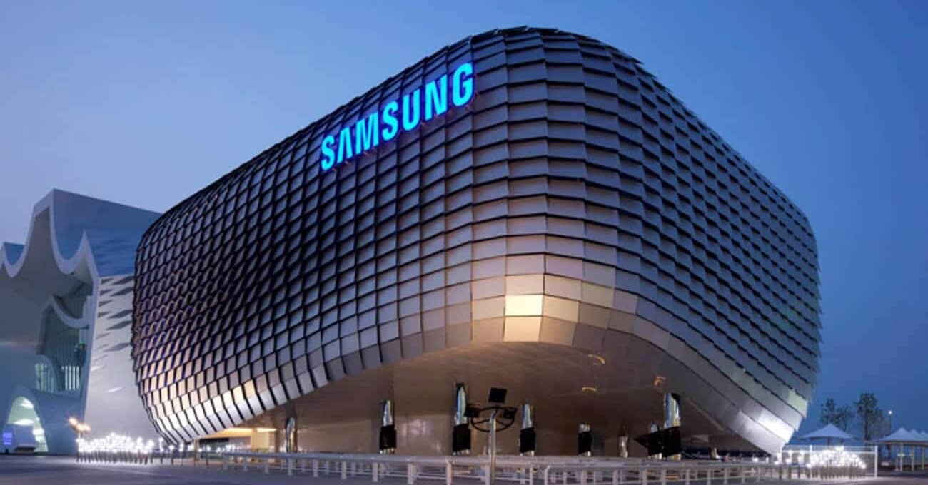 Fachada de prédio moderno revestida com módulos metálicos ondulados e iluminada à noite, com o logo azul "SAMSUNG" em letras luminosas no topo