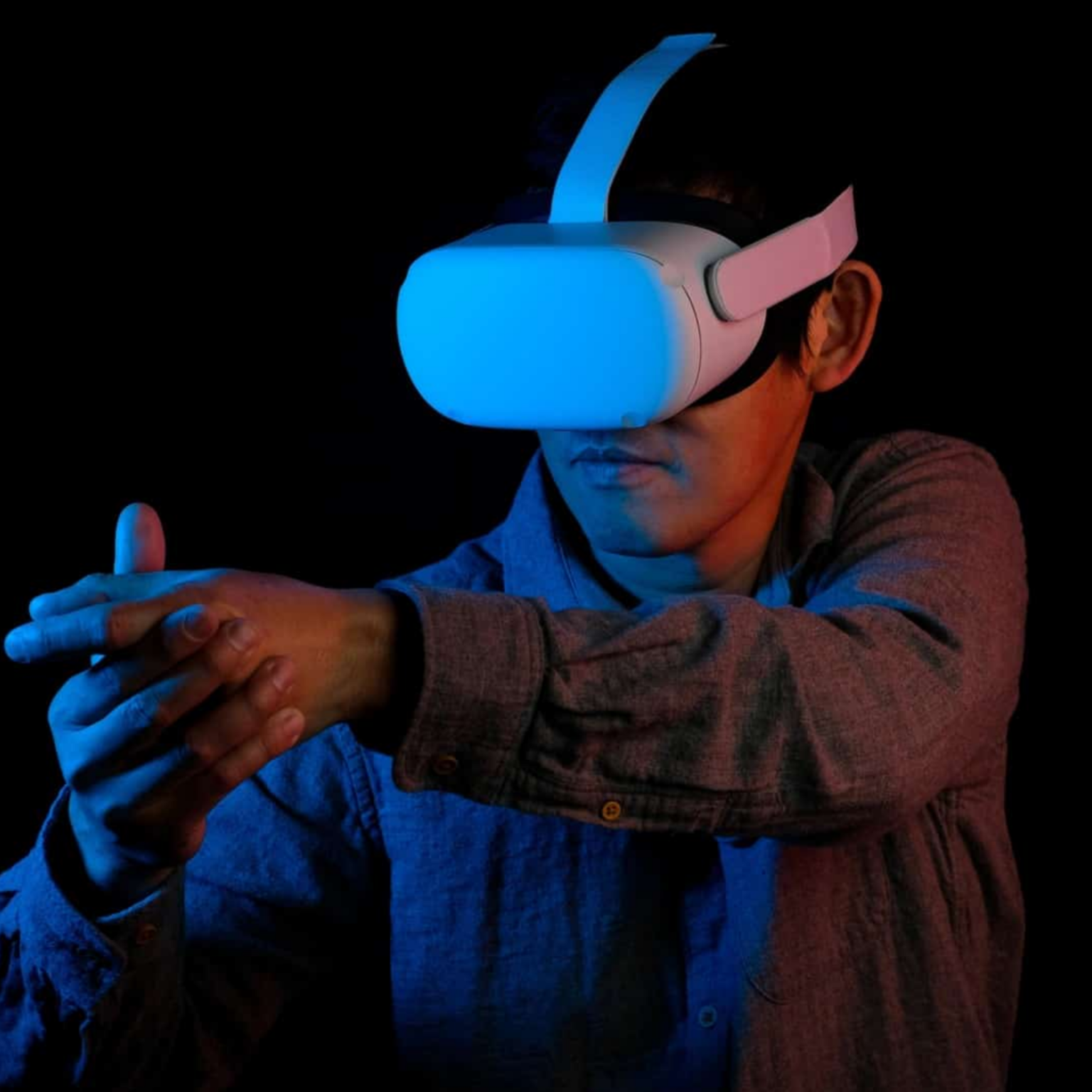 Homem de camisa xadrez usando óculos de realidade virtual branco e estendendo os braços em ambiente escuro iluminado por luz azul