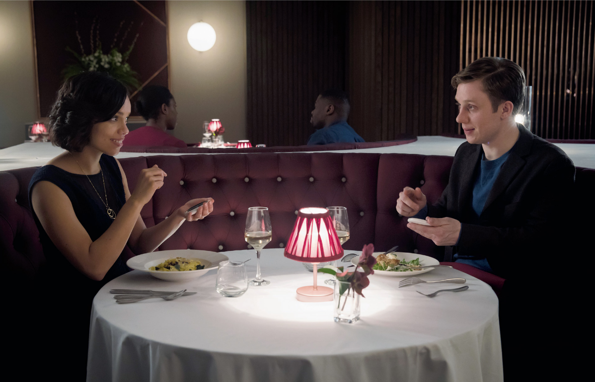 Casal jantando em restaurante à luz de abajur vermelho, segurando celulares, em cena que lembra o episódio "Hang the DJ" de Black Mirror