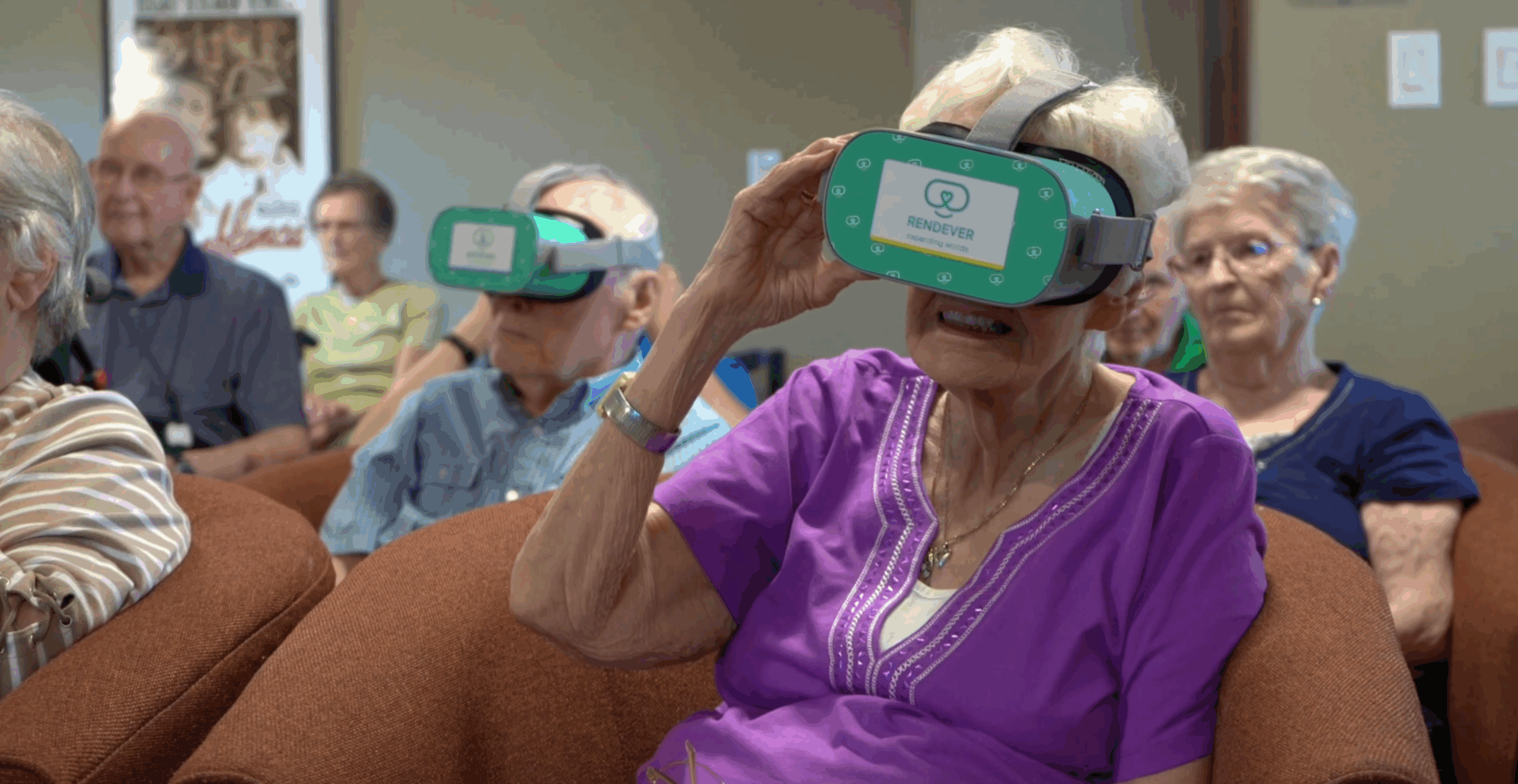 Idosos sentados usando óculos de realidade virtual verdes da marca Rendever, com uma senhora de blusa roxa ajustando o aparelho no rosto