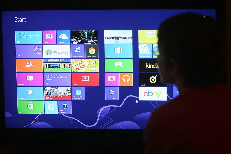 Tela inicial do Windows 8 em monitor grande, com blocos de aplicativos como Facebook, Chrome, eBay e Kindle, observada por uma pessoa de costas