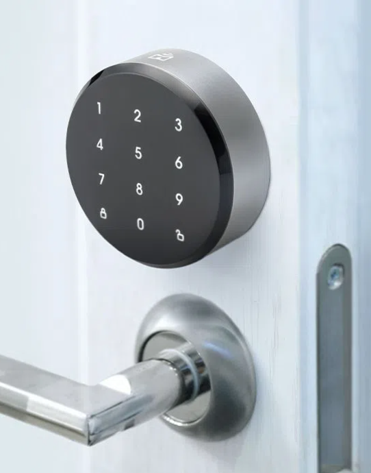 Fechadura inteligente prateada em porta branca, com teclado numérico touch circular preto com números de 0 a 9 e botões de trancar e destrancar