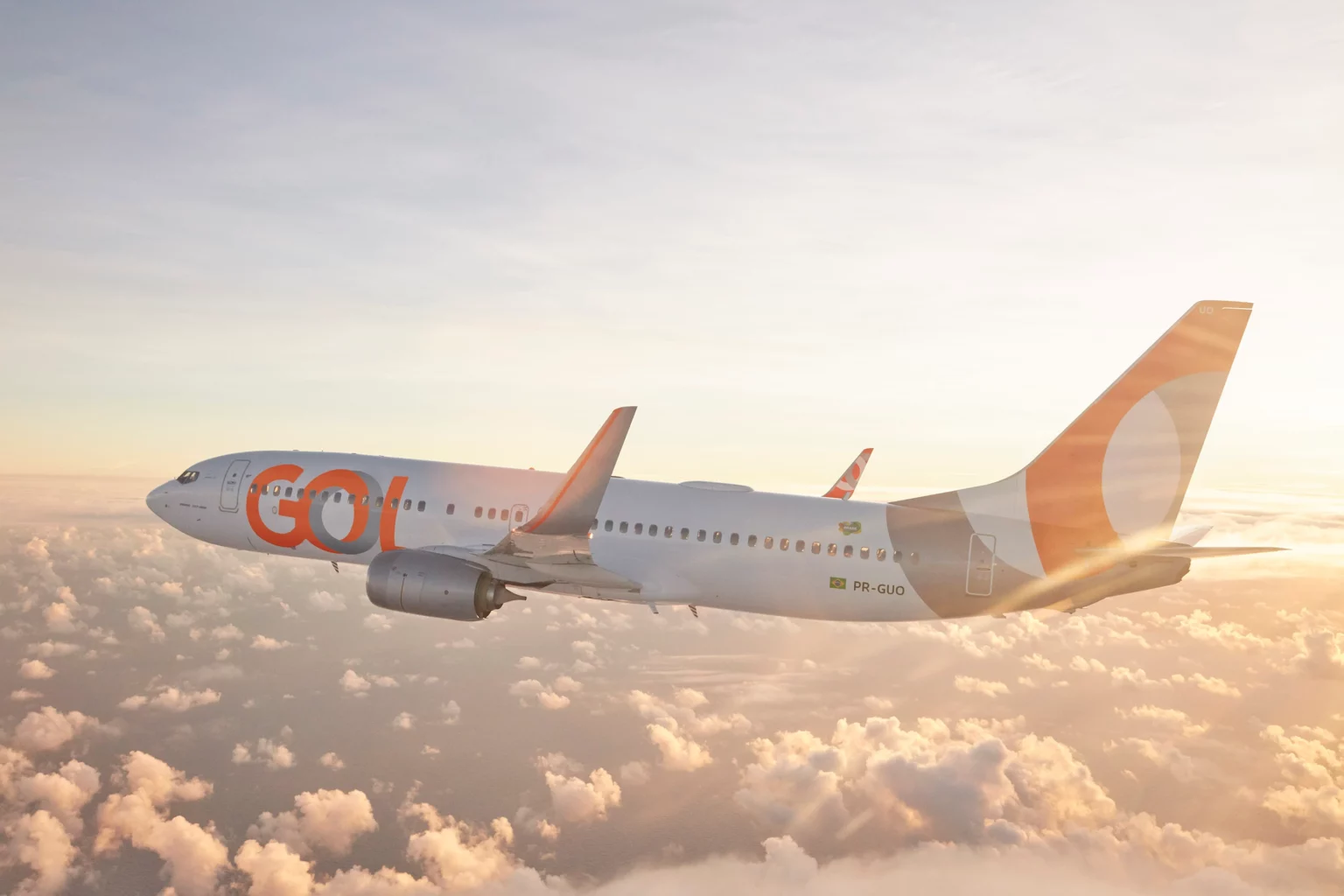 Avião da Gol com pintura branca e laranja voando sobre nuvens ao pôr do sol, com o logo GOL na fuselagem