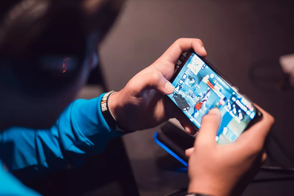 Pessoa segurando smartphone com as duas mãos e jogando um game de tiro em primeira pessoa na tela, em ambiente escuro