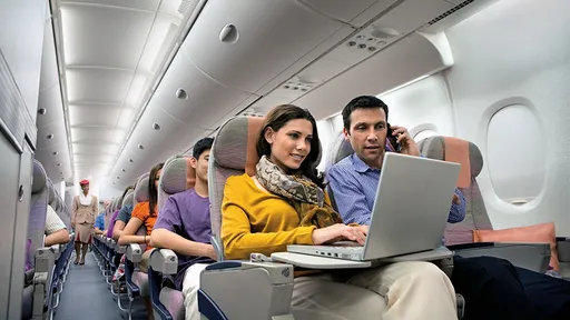 Passageiros sentados em cabine de avião, com notebook no colo e celular na orelha, enquanto comissária caminha pelo corredor