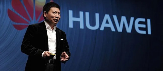 Executivo de terno escuro falando com microfone de cabeça em palco com logo da Huawei ao fundo