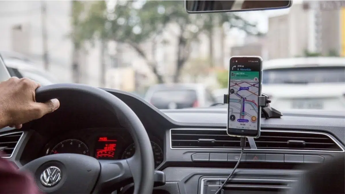 Motorista dirigindo carro Volkswagen com celular fixado no painel exibindo aplicativo de navegação com mapa e rota