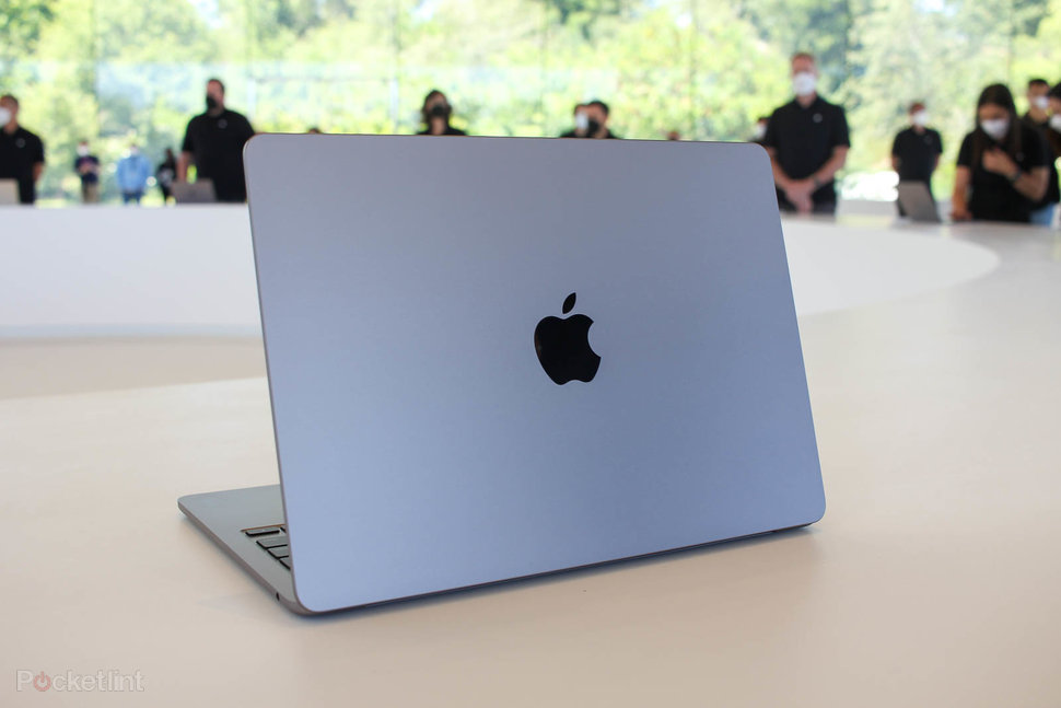 Notebook MacBook Air azul de costas sobre mesa branca, com o logo da Apple no centro da tampa e pessoas de camiseta preta ao fundo