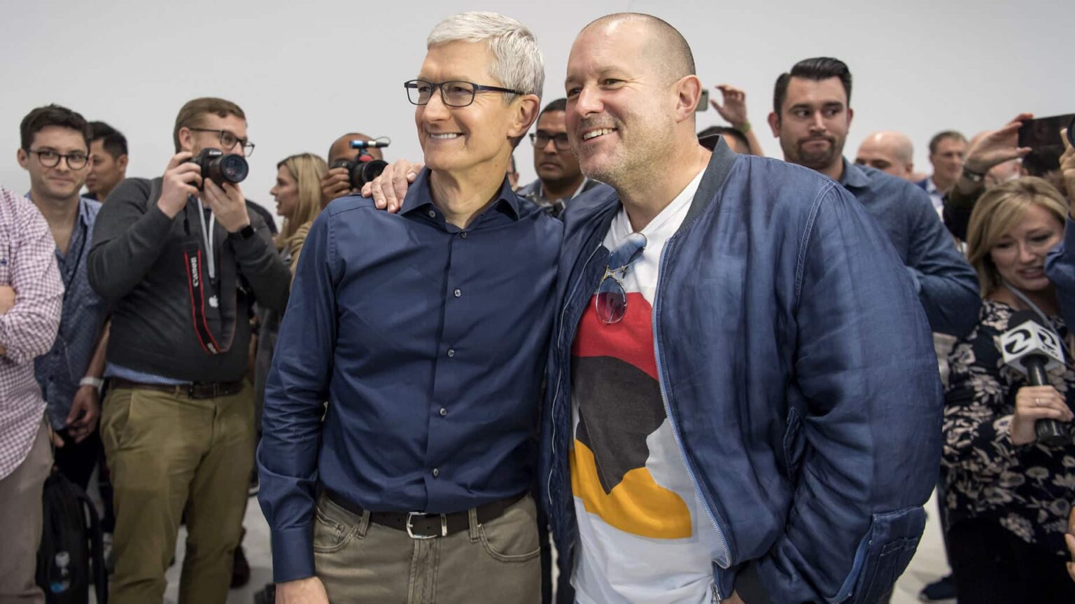 Dois homens sorrindo e posando lado a lado em evento com fotógrafos e jornalistas ao redor, um com camisa azul-marinho e outro com jaqueta jeans sobre camiseta…