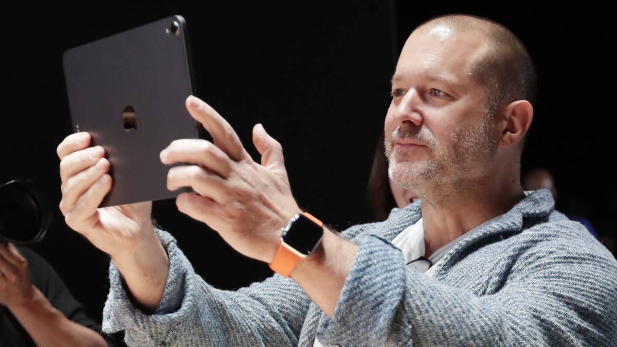 Homem de camisa cinza segurando um iPad cinza-escuro com as duas mãos, relógio Apple Watch com pulseira laranja no pulso, em ambiente escuro de evento