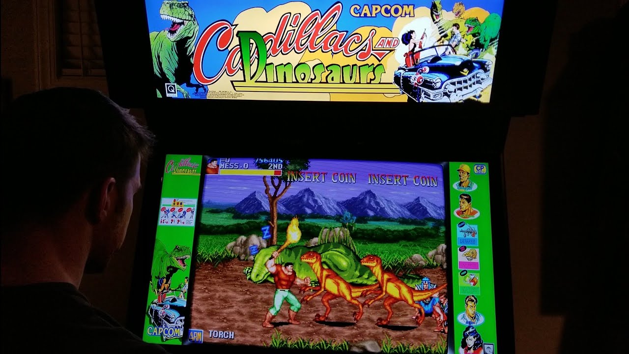 Pessoa jogando o fliperama Cadillacs and Dinosaurs da Capcom, com a tela mostrando um personagem enfrentando dinossauros em um cenário de montanhas