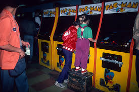 Duas crianças jogam em gabinetes de arcade Pac-Man em um fliperama, uma delas em pé sobre um caixote para alcançar a tela