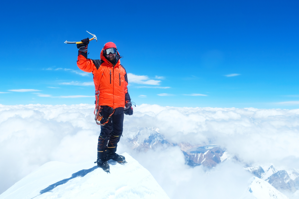 Alpinista de jaqueta laranja e capacete vermelho levanta picareta de gelo no topo de uma montanha nevada, acima de um mar de nuvens