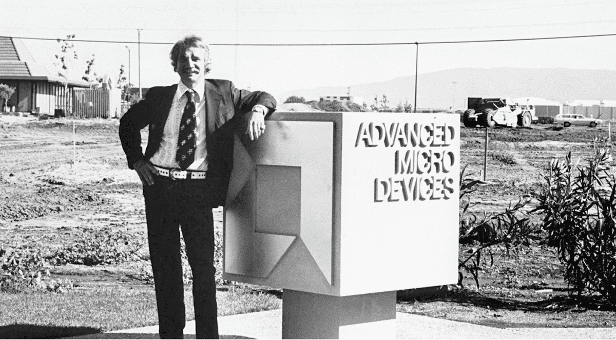 Homem de terno e gravata encostado em placa de concreto com o letreiro "Advanced Micro Devices", em terreno aberto, em foto em preto e branco