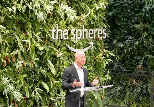 Homem de terno discursando em pódio de acrílico diante de parede verde com plantas, ao lado do letreiro "the spheres" com o logo da Amazon