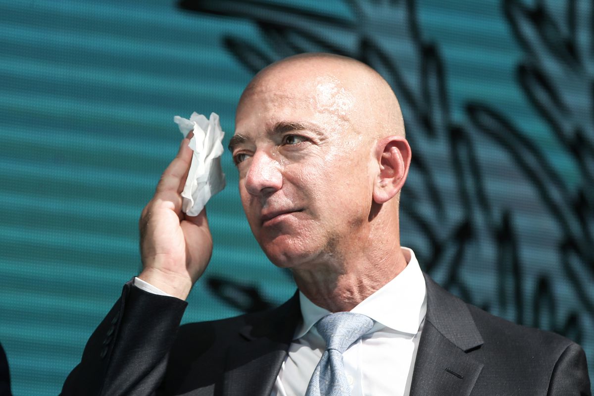 Homem careca de terno escuro e camisa clara, segurando um lenço de papel amassado perto do rosto, com fundo azul-turquesa