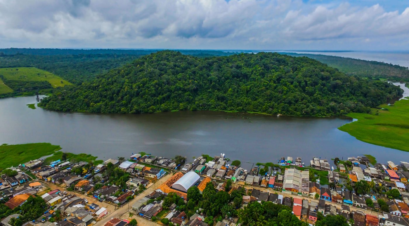Vista aérea de cidade ribeirinha às margens de rio largo, com floresta densa e morro coberto de mata ao fundo, sob céu nublado