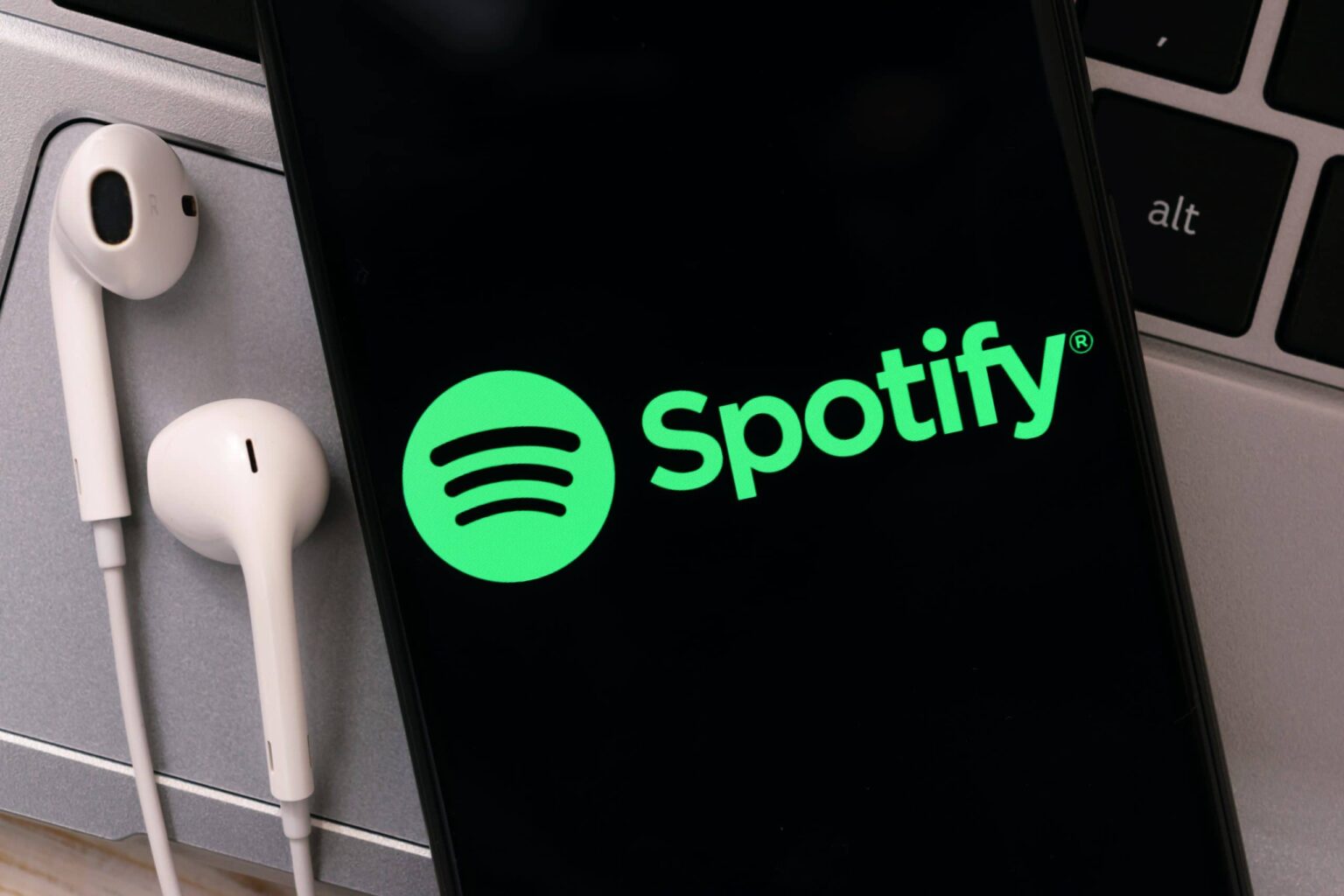 Smartphone com o logotipo verde do Spotify na tela escura, ao lado de fones de ouvido brancos sobre um teclado de notebook