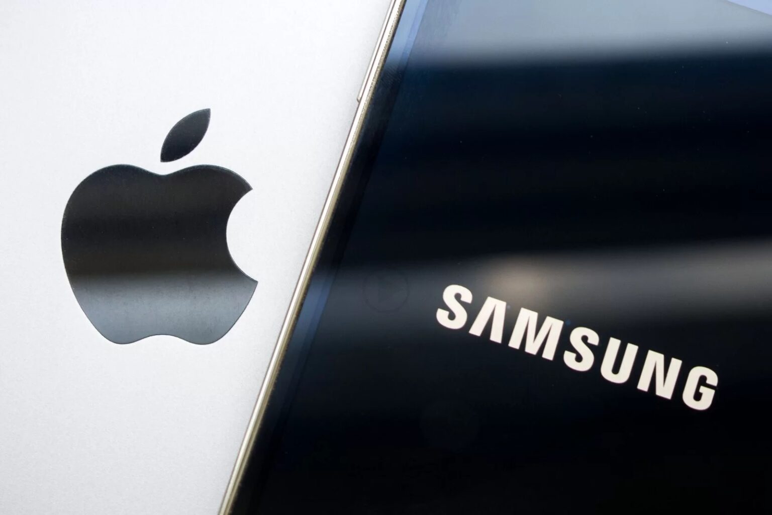 Logo da Apple em superfície prateada ao lado do logo da Samsung em painel preto, lado a lado em close