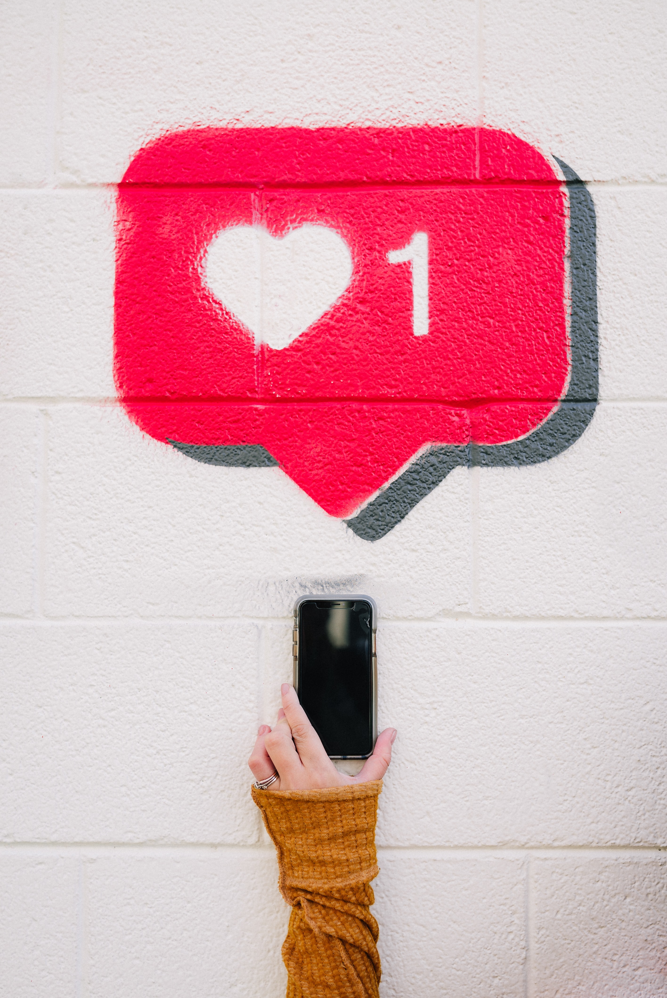 Mão segurando um smartphone em frente a uma parede branca com grafite de um balão vermelho de curtida com coração e o número 1