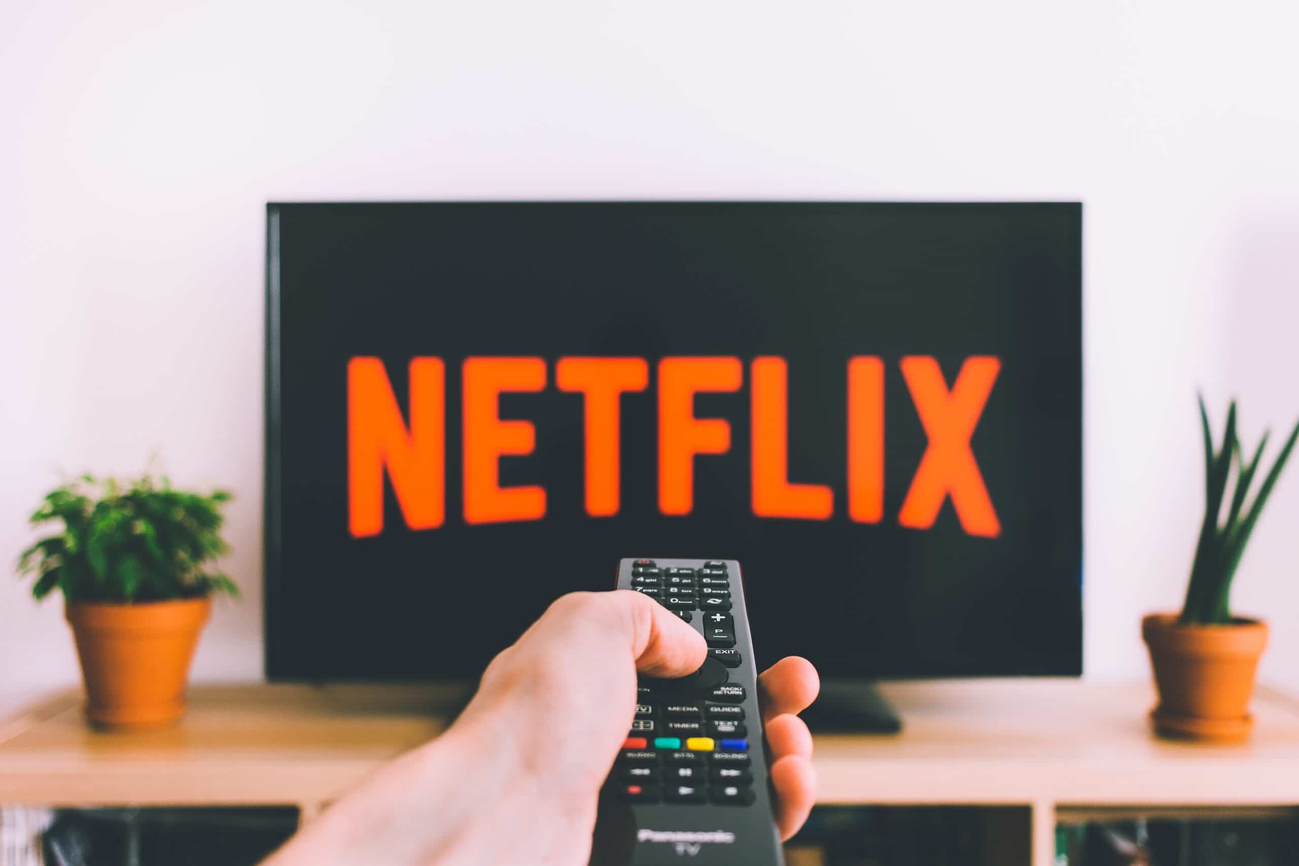 Mão segurando controle remoto apontado para TV exibindo o logo laranja da Netflix, com plantas em vasos nas laterais