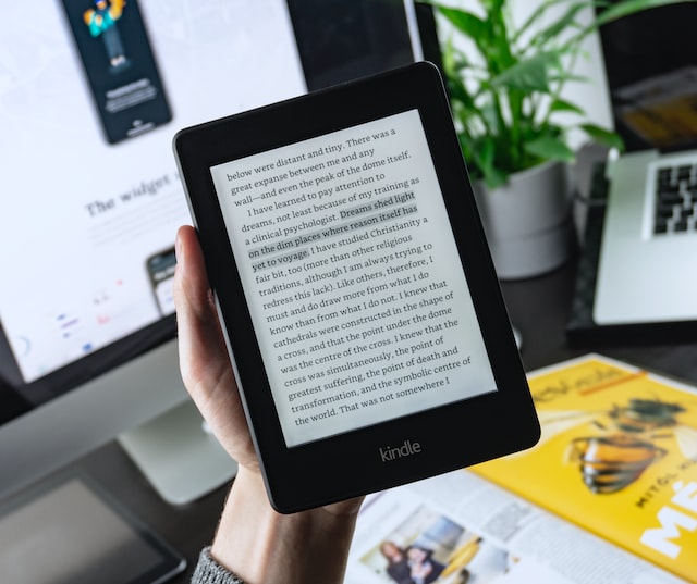 Leitor de e-books Kindle preto nas mãos de uma pessoa, exibindo um trecho de texto em inglês sobre um fundo de mesa com computador e plantas
