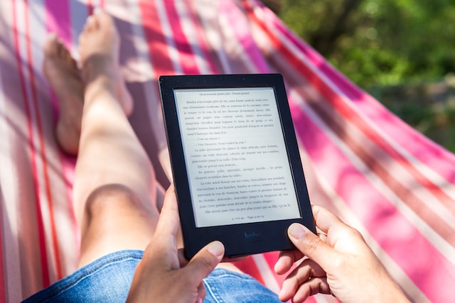 Pessoa lendo um e-reader preto em uma rede colorida ao ar livre, com texto em tela de tinta eletrônica