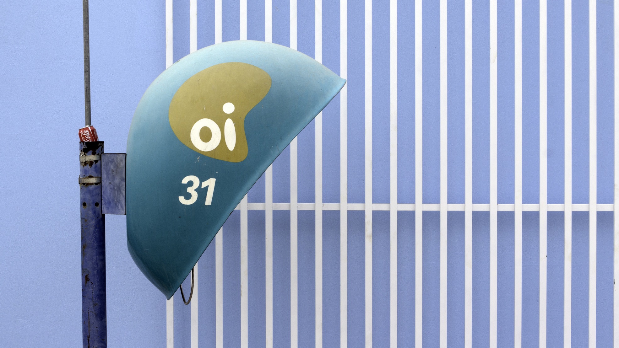 Orelhão azul-petróleo com o logotipo da Oi e o número 31, fixado em um poste diante de um muro de grades brancas e fundo lilás
