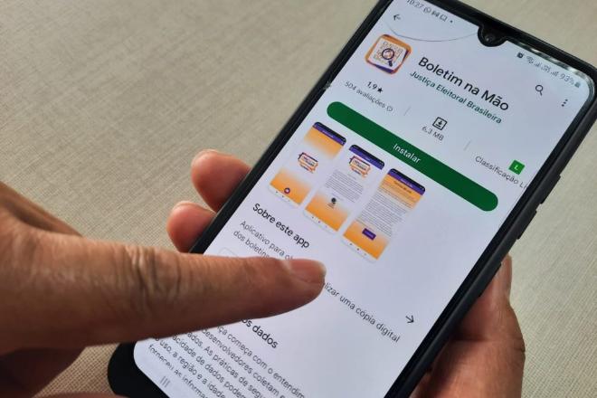 Mão segurando smartphone exibindo a página do app Boletim na Mão na Google Play, com botão Instalar e aviso sobre a cópia digital do boletim de urna