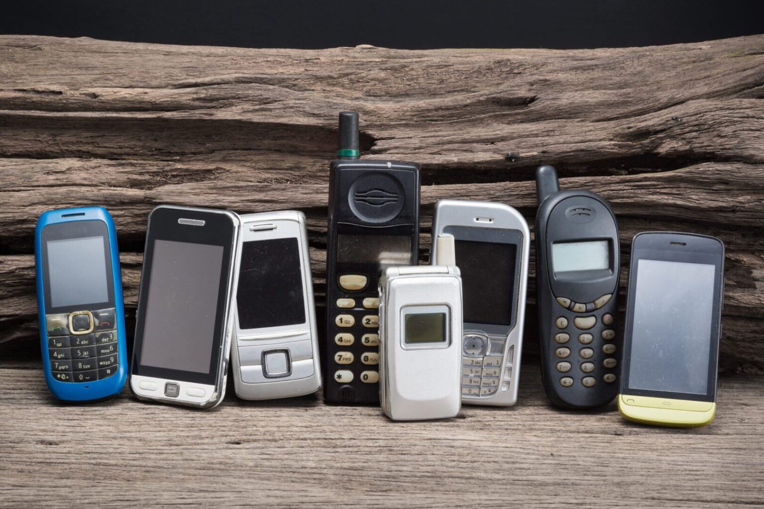 Sete celulares antigos de diferentes modelos e cores enfileirados sobre uma madeira envelhecida, incluindo aparelhos de teclado, de tampa e modelos de tela tou…