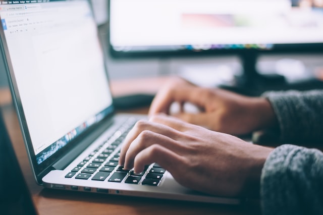 Mãos digitando em teclado de notebook prateado, com um monitor de computador desfocado ao fundo sobre uma mesa de madeira
