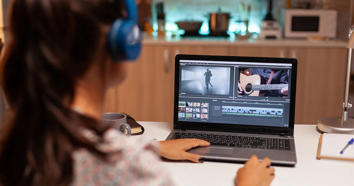 Pessoa de costas usando headphones e editando vídeo em um notebook, com timeline e uma cena de violão na tela, em cozinha iluminada