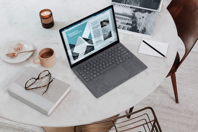Notebook prateado sobre mesa de mármore branca, ao lado de óculos, caneca, colher e vela, com tela exibindo página de arquitetura geométrica