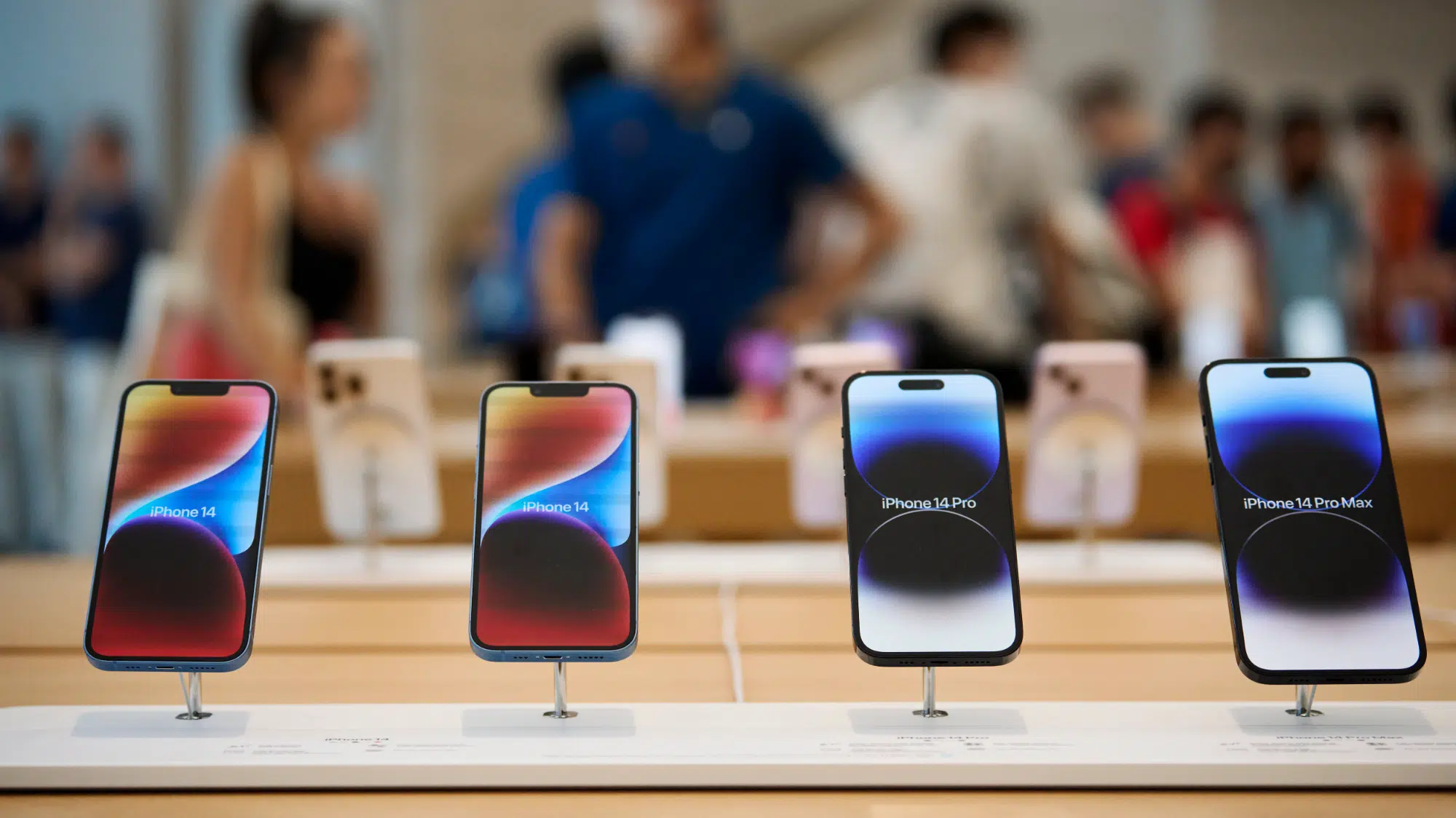 Quatro iPhones 14, 14 Plus, 14 Pro e 14 Pro Max expostos em loja Apple, com pessoas desfocadas ao fundo