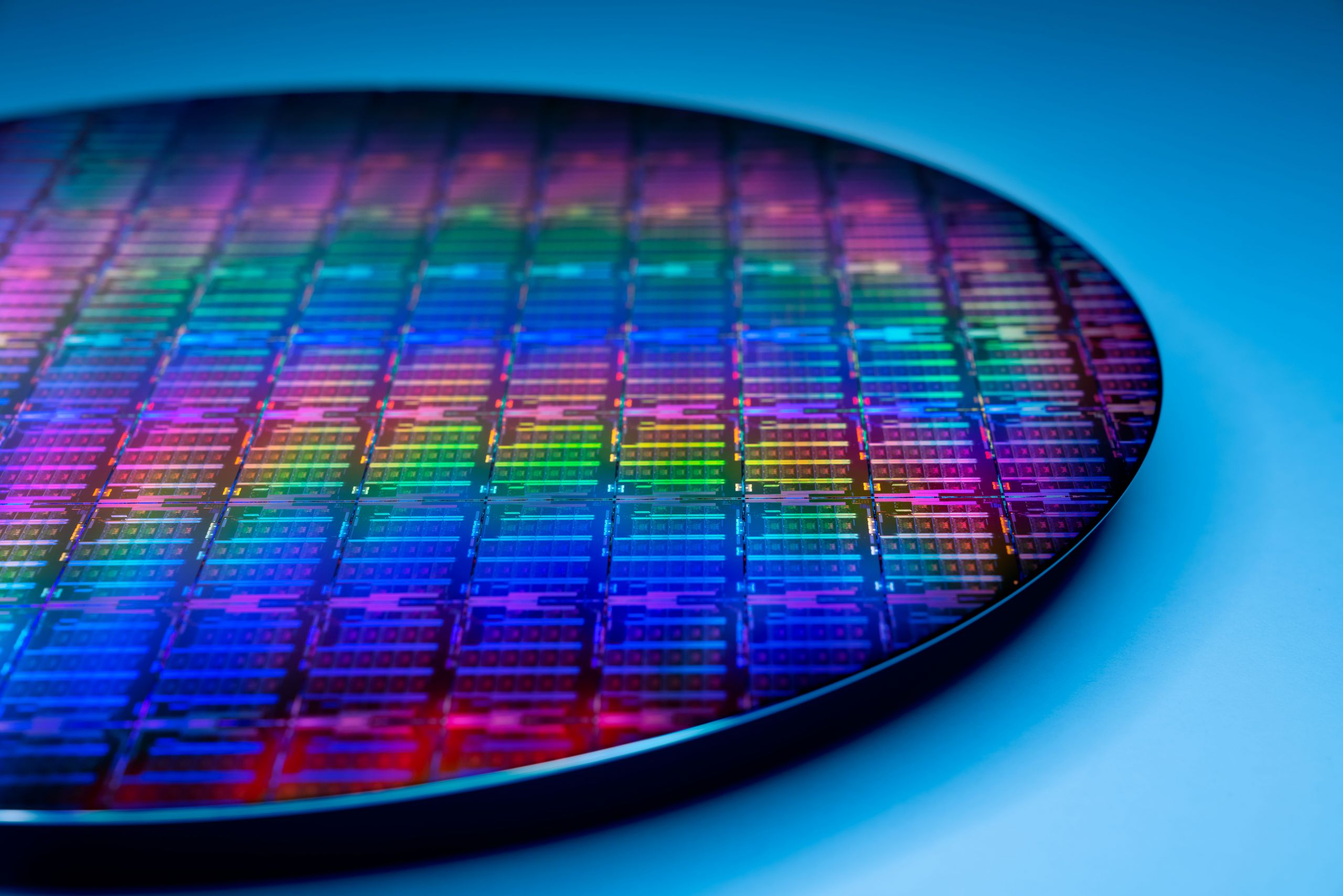 Wafer de silício com chips em tons iridescentes de azul, roxo, verde e vermelho sobre fundo azul, mostrando a matriz de processadores